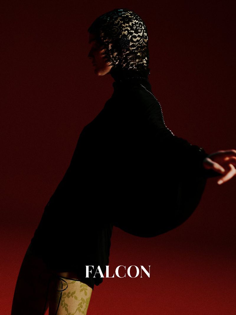 Fashion editorial shot of a woman in black clothing and a lace head covering posing in a studio with a red background.