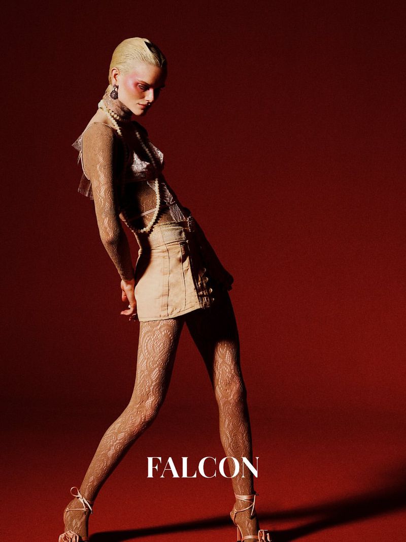 Fashionable woman in a pose with lace tights, a beige skirt, and a pearl necklace against a red background.