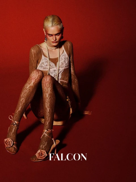 Adult woman poses indoors against a red backdrop wearing a lace top, jewelry, lace stockings, and floral high heels.