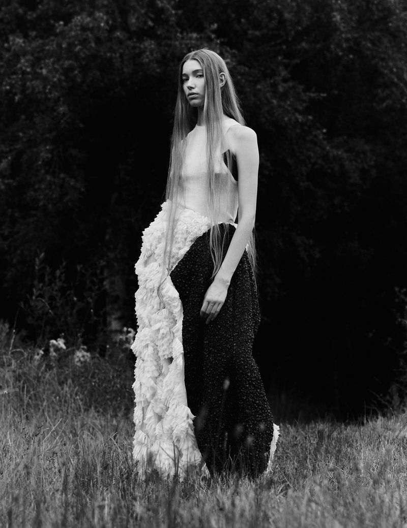 Monochromatic portrait of a solitary female figure in a field, draped in an avant-garde white and black textured dress, against a dark, blurred backdrop.