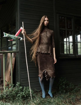 Adult female posing outdoors in a brown textured midi dress with decorative trim and blue hosiery, holding a white pole with fish kites.