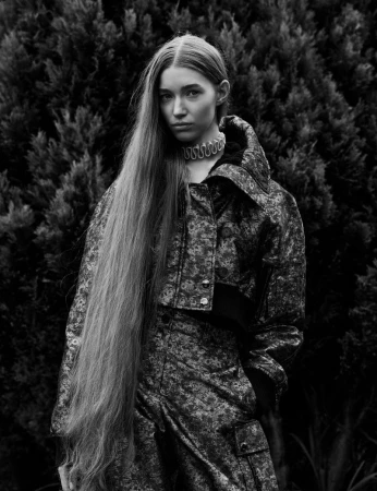 Black and white portrait of a female model with long hair wearing a patterned jacket outdoors against dense foliage.