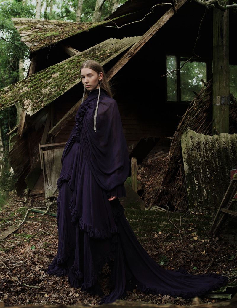 Woman in a long draped dress poses in front of an old, mossy cabin surrounded by trees in an outdoor setting with a medium shot.