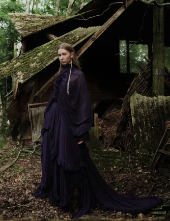 Woman in a long draped dress poses in front of an old, mossy cabin surrounded by trees in an outdoor setting with a medium shot.