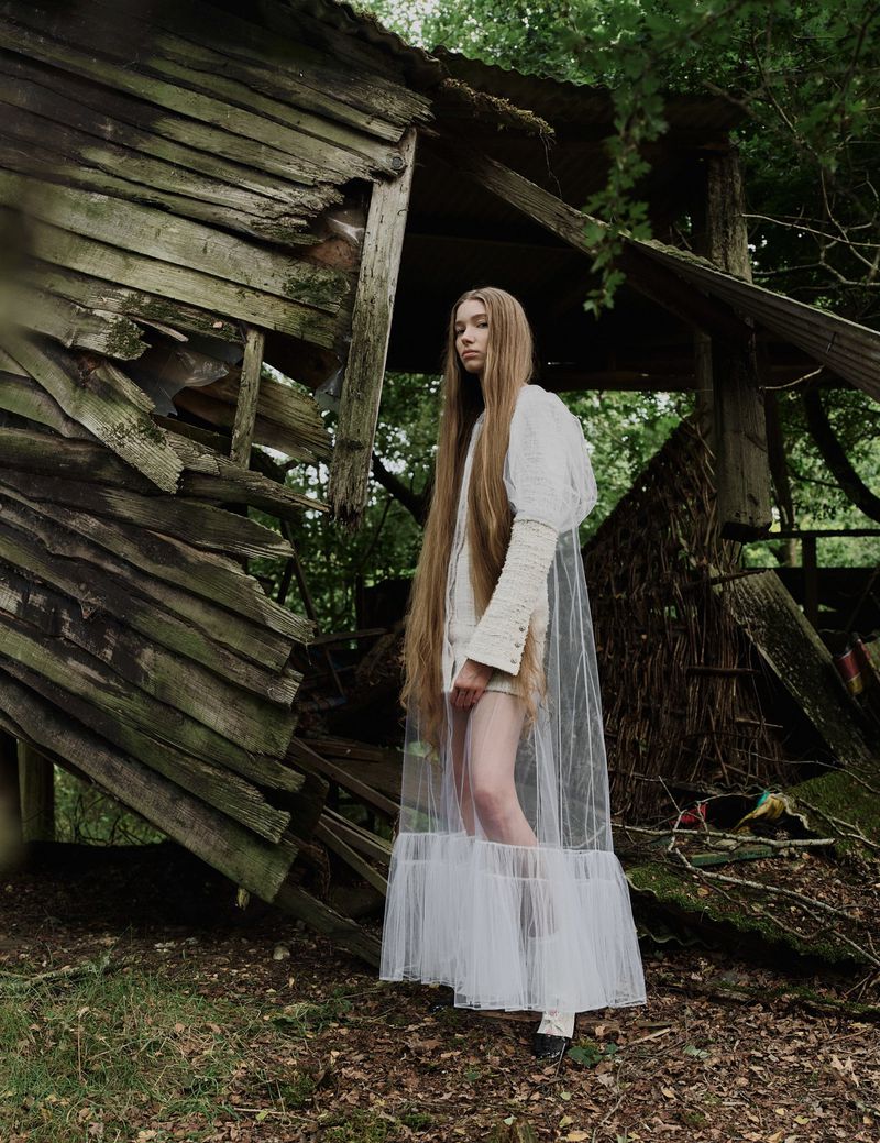 A woman with long hair stands in front of a dilapidated wooden structure in an overgrown outdoor setting, creating a blend of fashion and decay.