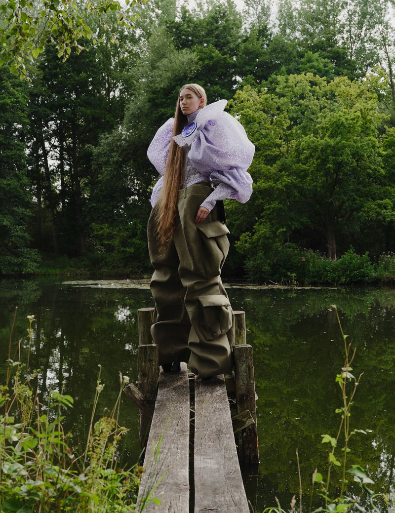 A woman in statement fashion stands on a wooden structure above a body of water, surrounded by lush greenery and trees on an overcast day.