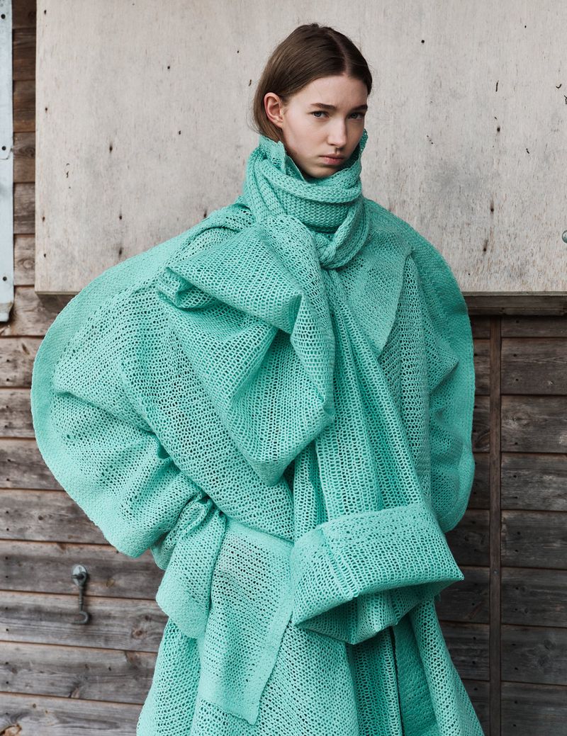 Fashion shot of a young woman wearing an oversized aqua, woven coat, posing in front of a weathered wooden wall in daylight.