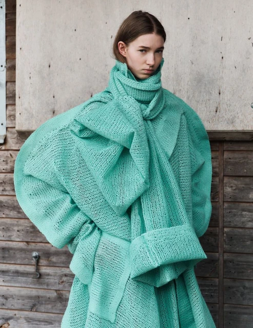Fashion shot of a young woman wearing an oversized aqua, woven coat, posing in front of a weathered wooden wall in daylight.
