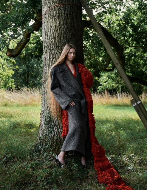 An adult woman stands near a tree wearing a gray wool coat with a belt and long hair. She is outdoors in a grassy area.