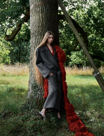 An adult woman stands near a tree wearing a gray wool coat with a belt and long hair. She is outdoors in a grassy area.