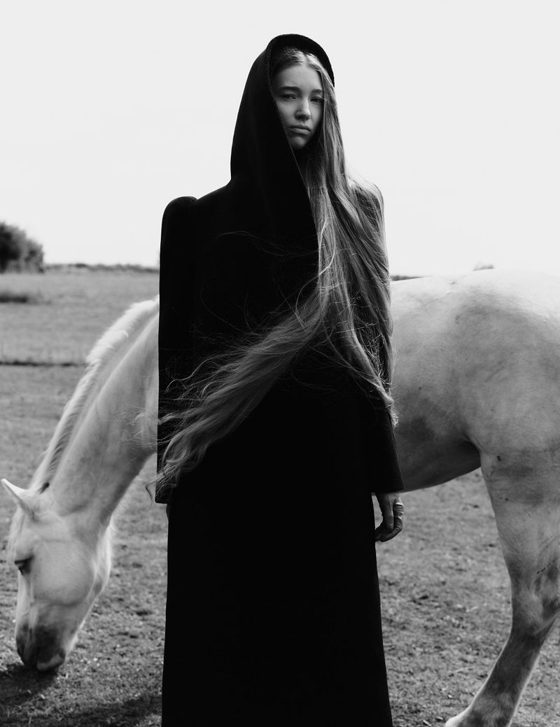 Monochrome image of a young woman in a hooded cloak standing next to a white horse in a grassy field under an overcast sky.