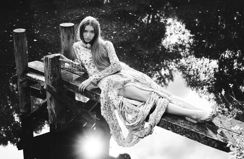 A woman in a floral dress lies barefoot on a wooden dock over a dark body of water reflecting the sky in a black and white outdoor shot.