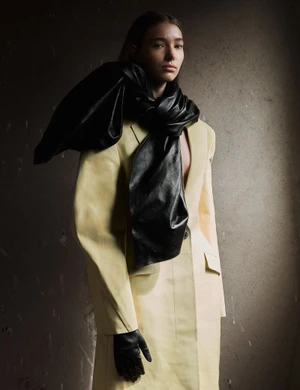 Model Liz Schweikl wearing a pale yellow tailored coat, large black leather scarf, and black leather glove against a textured wall.