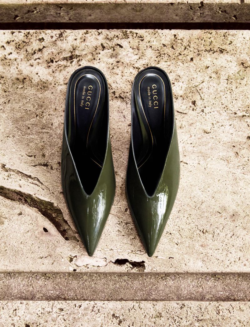 Olive green patent leather Gucci pointed-toe mule shoes on a textured marble surface, showcasing elegant footwear and luxury branding.