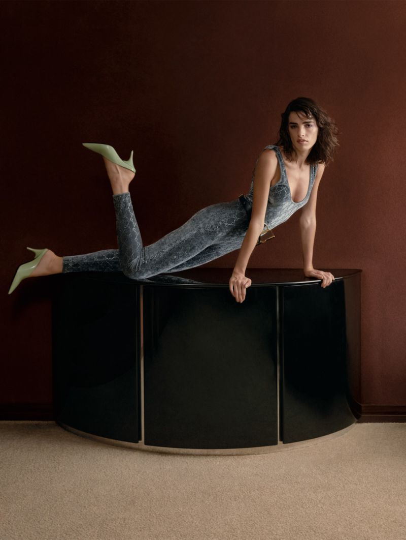Woman in a gray jumpsuit and green heels poses on a black cabinet against a warm brown wall in a fashion editorial shot.