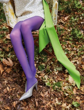 Model seated outdoors in an armchair wearing purple tights and grey heels amidst fallen leaves.