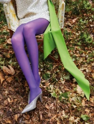 Model seated outdoors in an armchair wearing purple tights and grey heels amidst fallen leaves.