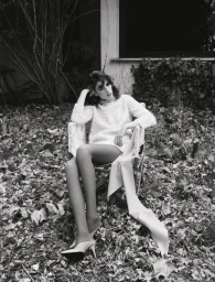 Black and white portrait of a woman seated in a folding chair outdoors among fallen leaves, wearing a patterned top and heels.