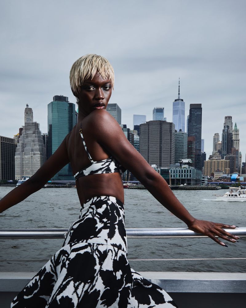 Fashionable woman in matching bikini top and skirt poses in front of cityscape by the water on an overcast day.