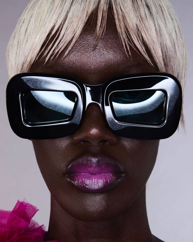 Close-up portrait of an adult female model with dark skin and short, blonde hair wearing oversized, black sunglasses and pink lipstick.