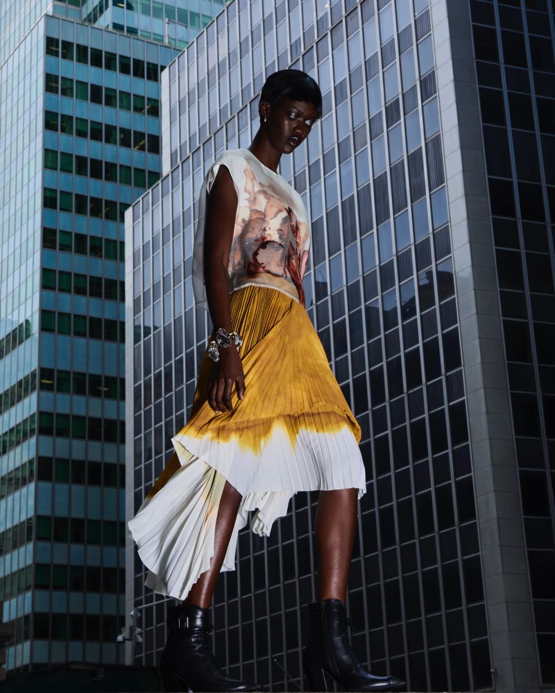 Fashion editorial featuring a Black woman wearing a yellow pleated skirt and top in front of modern glass and steel skyscrapers.