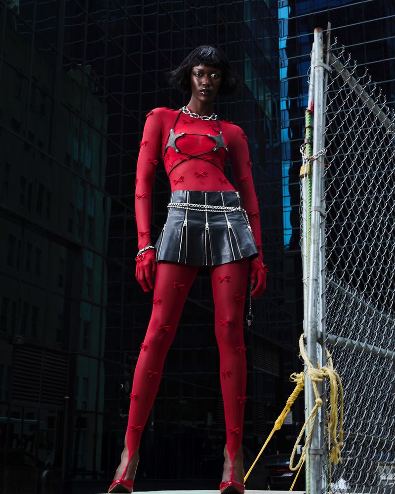 Model wearing a red sheer patterned top, black leather miniskirt, and red tights against an urban background with a chain-link fence.