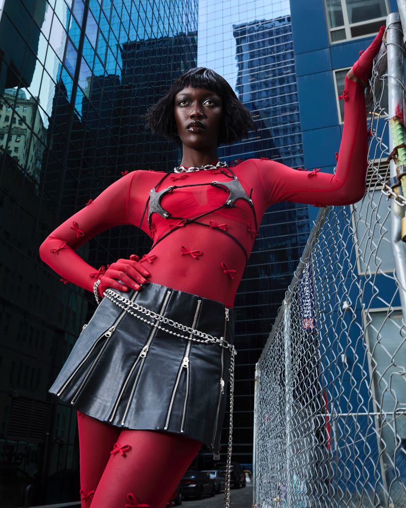 Fashionable Black woman in red bodysuit and black mini skirt poses in front of glass skyscrapers and chain-link fence in urban setting.