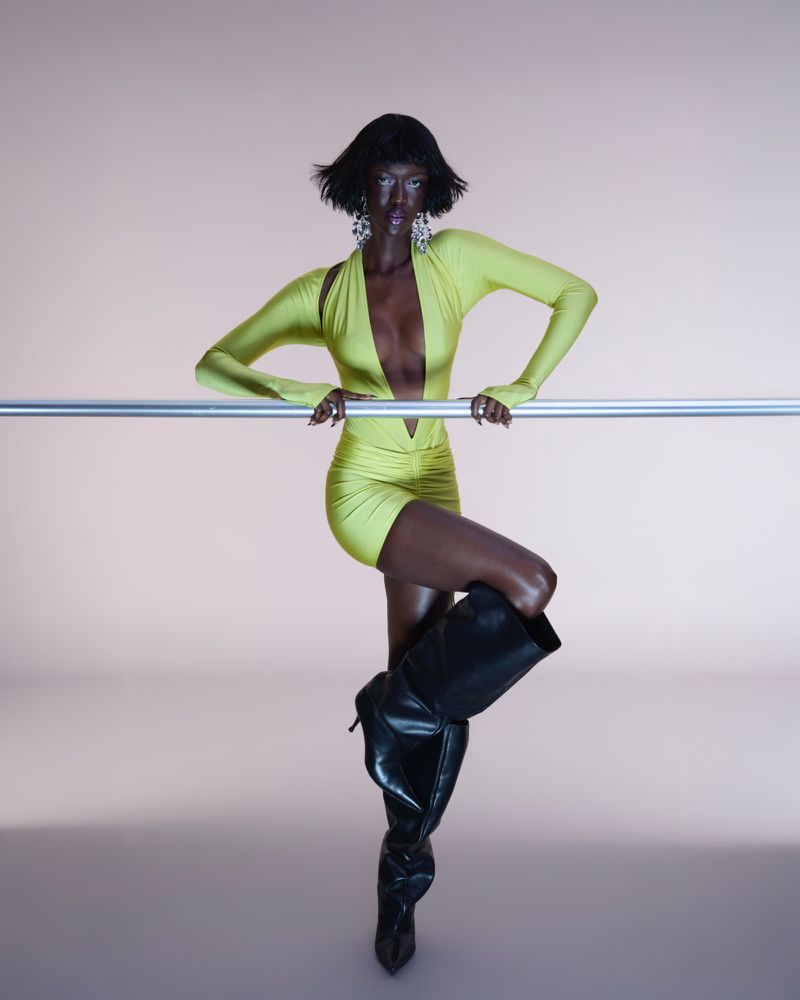 Elegant Black woman in a neon yellow dress poses against a light pink backdrop, leaning on a metal bar and wearing black high-heeled boots.
