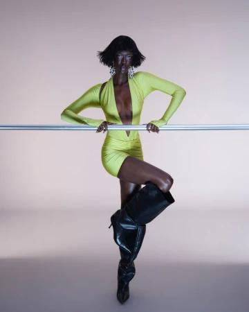 Elegant Black woman in a neon yellow dress poses against a light pink backdrop, leaning on a metal bar and wearing black high-heeled boots.