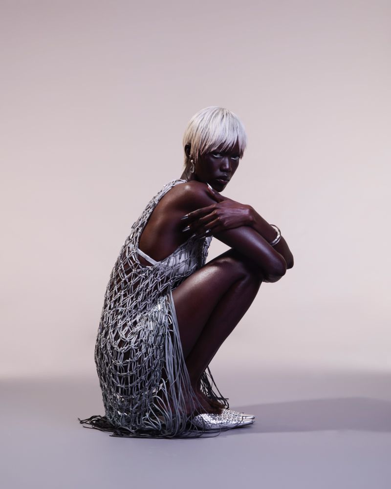 Woman with dark skin and white hair in a fringed silver dress squatting indoors against a light background, looking directly at the camera.