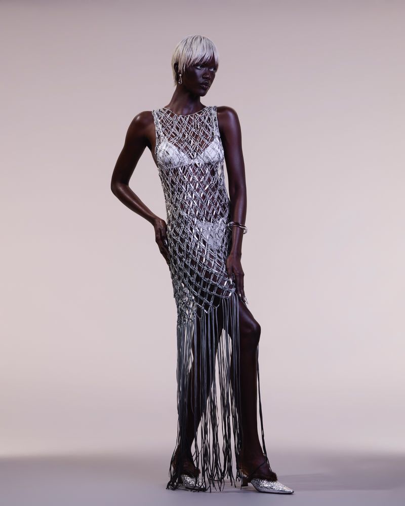 A slender dark-skinned woman with platinum hair poses in a silver metallic fringe dress and matching shoes against a neutral background.