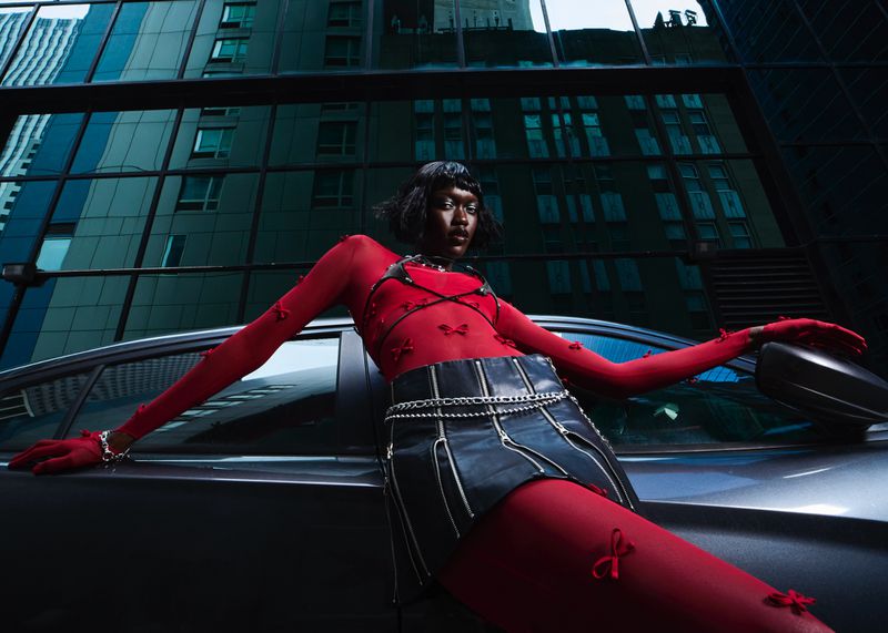 A dark-skinned woman in a red mesh top and black mini-skirt poses against a car with city buildings in the background.