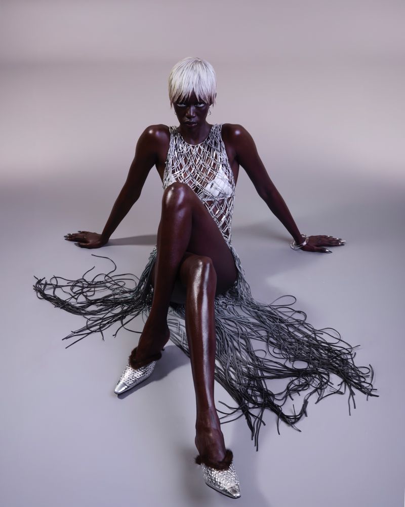 Fashion portrait of a Black woman in a silver dress with a fringe skirt sitting on a grey floor, wearing silver heels in a studio setting.