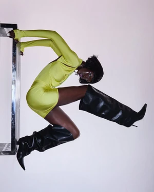 Model Adama Jobe posing dynamically in a chartreuse mini dress and black leather slouch boots against a white studio background.