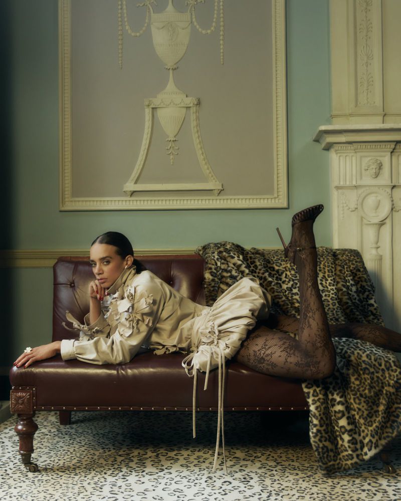 A woman in a beige jacket and dress poses on a brown leather couch with patterned stockings and leopard-print blanket in an indoor setting.
