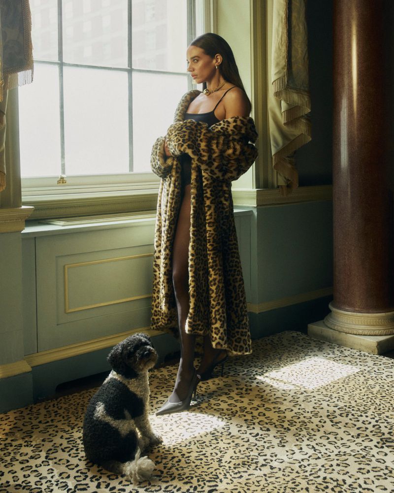 Woman in animal-print coat poses with dog in leopard-print room near window; fashion editorial, luxury lifestyle, animal print on animal print.