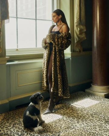 Woman in animal-print coat poses with dog in leopard-print room near window; fashion editorial, luxury lifestyle, animal print on animal print.