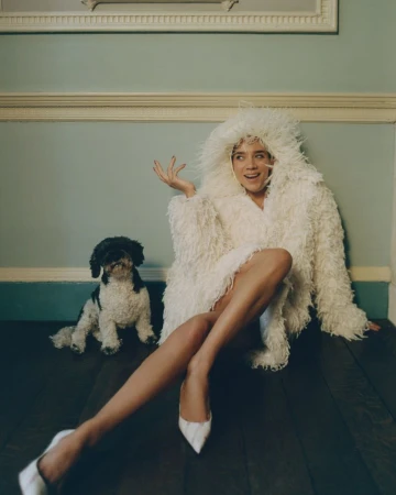 Woman in a furry white hooded coat poses indoors on a wood floor with a dog. She wears white heels in a portrait-style shot.