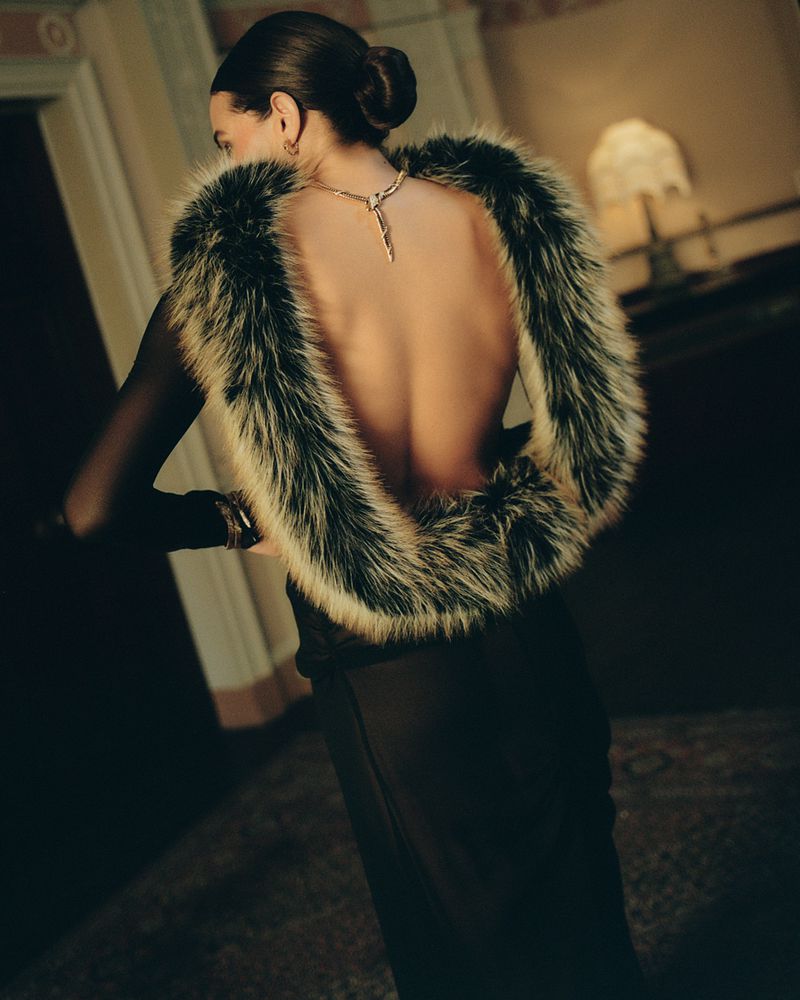 Back view of a woman in long black dress and fur stole, wearing jewelry indoors with warm lighting and neutral color palette.