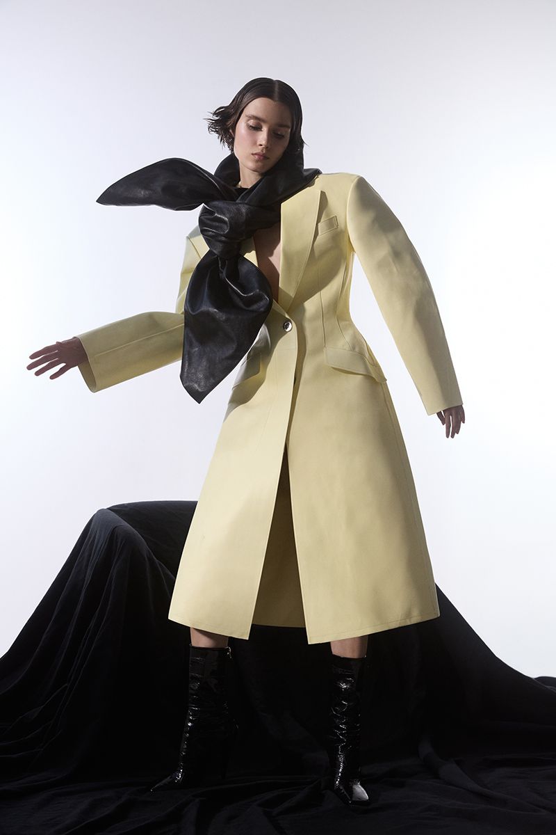 Fashion model posing in a pale yellow coat with a large black bow, patent leather boots, standing against a black drape on a white background.