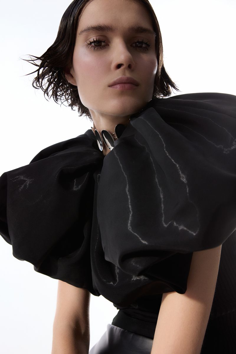 Close-up of a fashion model wearing a black top with large, puffed sleeves and a silver collar, against a bright, neutral background.