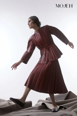 Fashion model poses in a pleated burgundy jacket and skirt by MOJEH against a neutral backdrop and flooring in a studio shot.