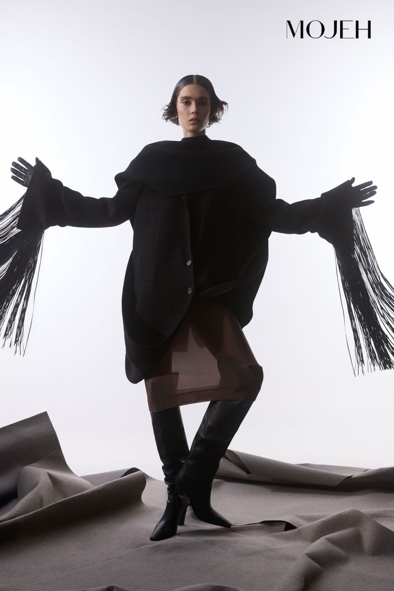 Fashion editorial shot of a woman in a black coat with fringe sleeves and black boots, posing in a studio with a grey fabric background.