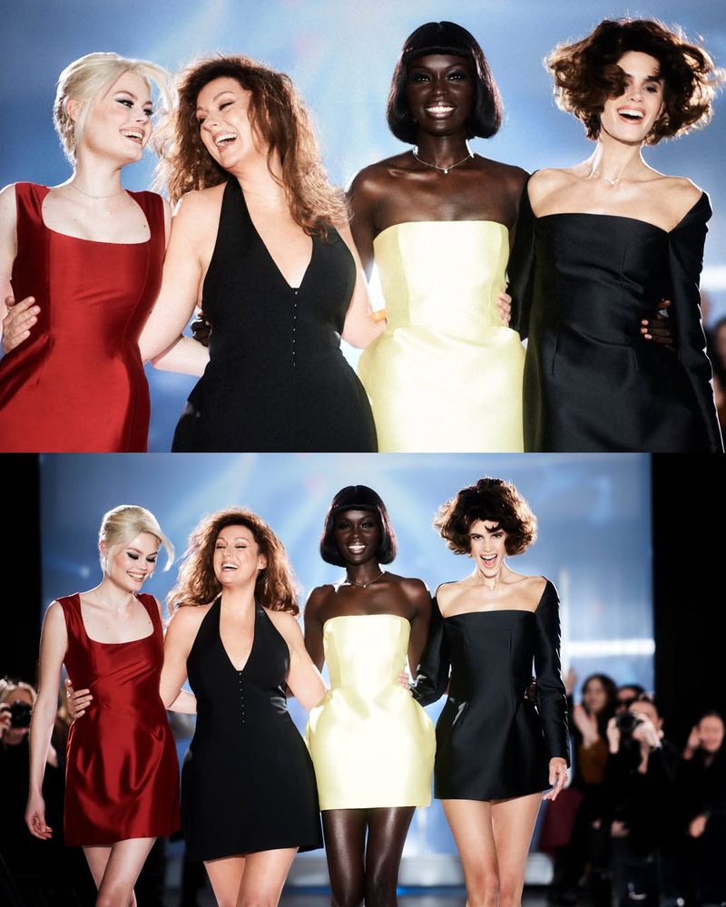 Four smiling female fashion models walk arm in arm down a runway in black, yellow, and red dresses during a fashion show.