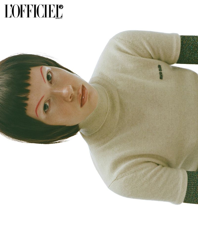Fashion editorial portrait of a young fair-skinned woman with dark blunt bangs, wearing a beige turtleneck sweater against a white background.