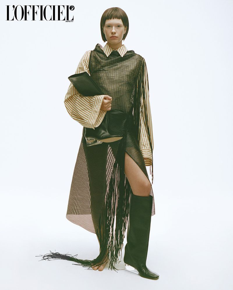 Fashion editorial shot: Brunette model in striped blouse, mesh fringed dress, and boots, holding a black document case against a white backdrop.