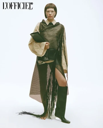 Fashion editorial shot: Brunette model in striped blouse, mesh fringed dress, and boots, holding a black document case against a white backdrop.