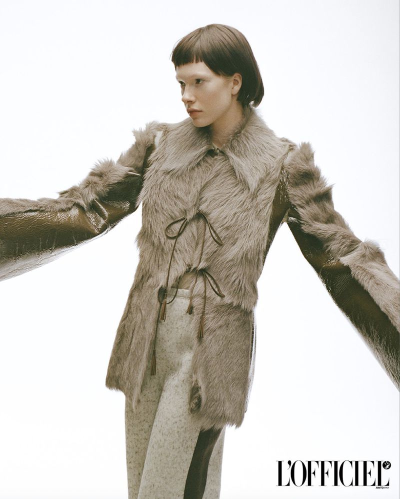 A pale-skinned woman poses in a faux fur vest with a glossy brown jacket and gray pants against a bright white backdrop, fashion editorial.