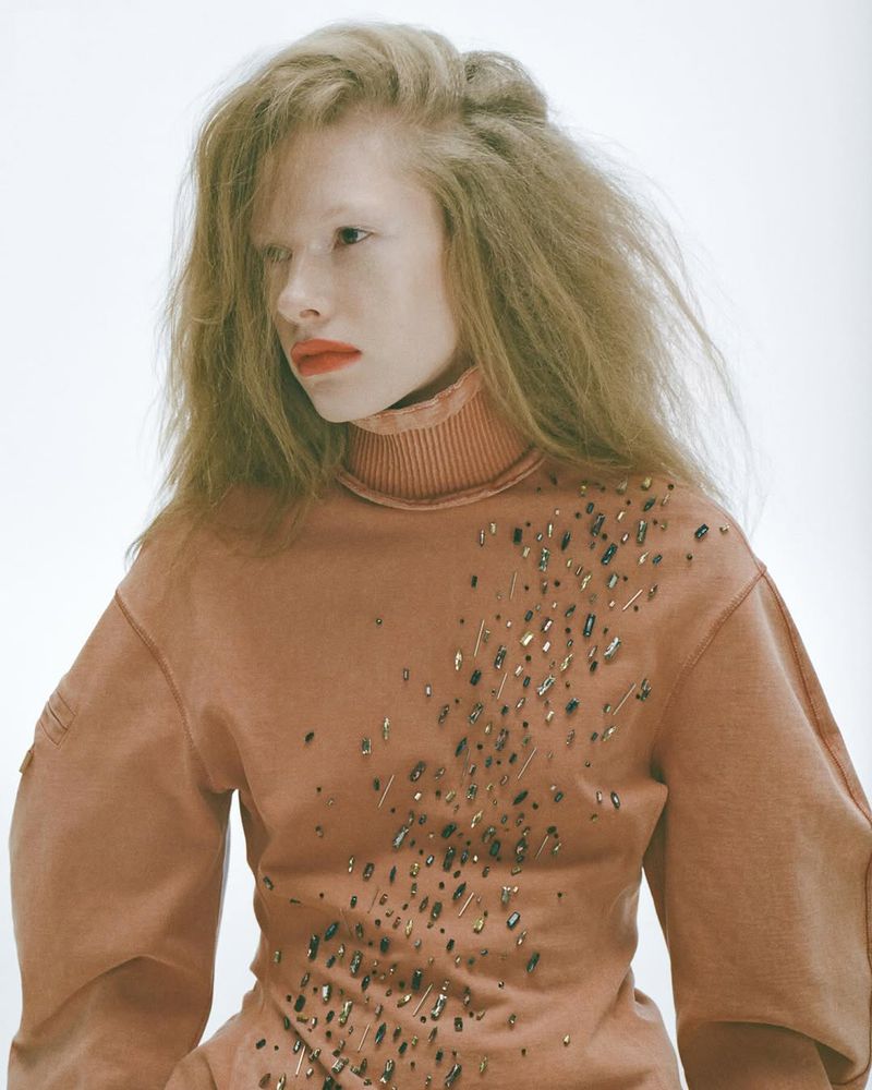 Close-up studio portrait of a fashion model with blond, voluminous hair, wearing a turtle neck sweatshirt with decorative accents.