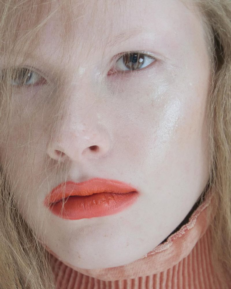 Close-up portrait of an albino woman with fair skin and blonde hair, showcasing coral lipstick and a high-neck ribbed sweater.